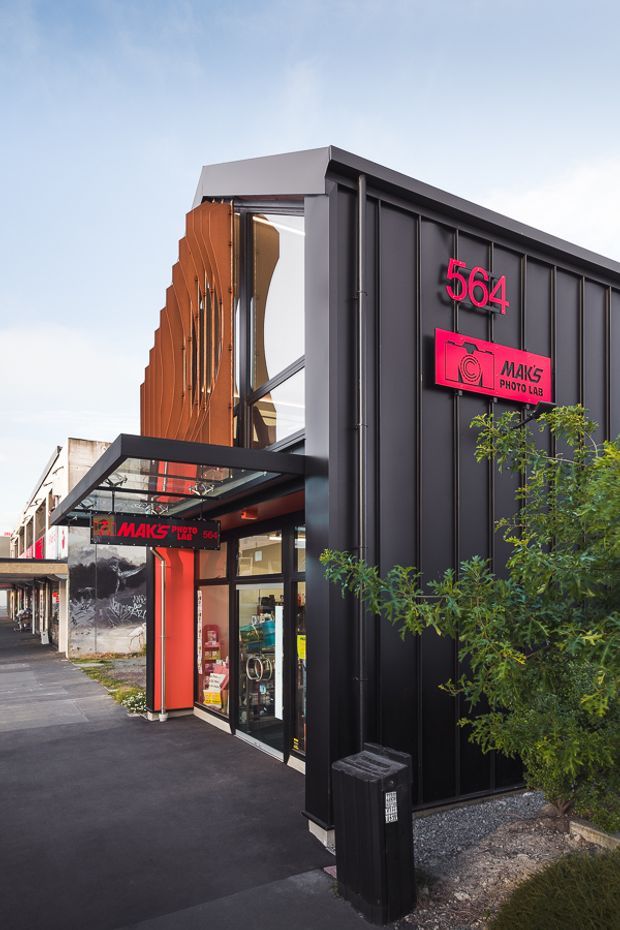 Street-level exterior view of a contemporary commercial photo lab at 564, with a bold black vertically ribbed metal facade, bright pink signage and entry canopy. The building has large glazed openings, orange-toned vertical facade elements, and sits along a paved urban retail frontage with planted landscaping.
