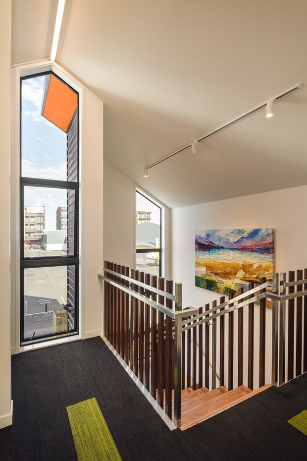 Bright contemporary stair landing with a switchback staircase enclosed by dark timber vertical balustrades and silver metal handrails. The landing has dark carpet with lime-green accent strips, white walls and ceiling, tall black-framed windows, track lighting, and a colourful abstract artwork. Urban buildings and sky are visible outside the glazing.