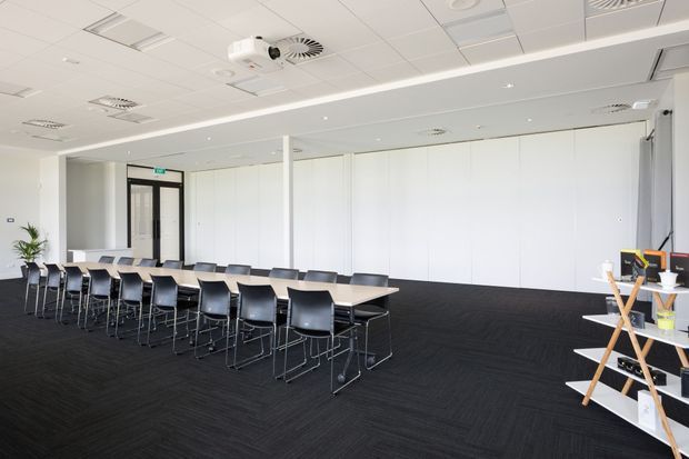 Bright, spacious commercial meeting or training room with long rows of rectangular tables and black chairs. The room has white folding or operable wall panels, a suspended acoustic tile ceiling with recessed lighting and HVAC fixtures, dark carpet flooring, a ceiling-mounted projector, and a refreshment or display stand at the right. Glazed double doors with an exit sign are visible at the rear.