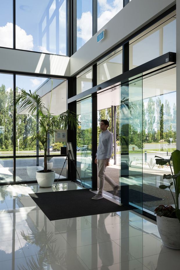 A bright, double-height modern entry lobby with extensive floor-to-ceiling glazing and black-framed glass doors. A person is entering through the automatic sliding entrance, while potted palms, a dark entrance mat, glossy light floor tiles, and strong reflections define the space. The lobby opens visually to landscaped grounds outside and receives abundant natural light through the glazed facade and overhead clerestory-like windows.