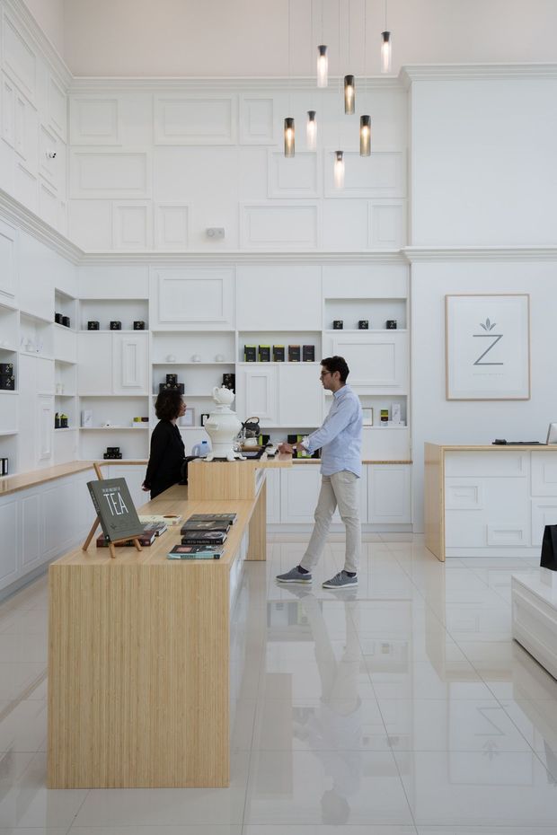 Bright, refined retail or commercial interior with a high ceiling, extensive white decorative wall panelling and built-in display cabinetry, glossy white tiled flooring, and a large light-wood island counter. Pendant lights hang above the central workspace, while shelving displays small products and books. Two people stand near the island. The space has a clean, contemporary-classic appearance.