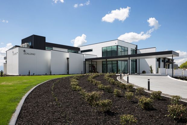 Exterior view of a contemporary commercial building with a white and dark charcoal facade, extensive glazed entry and office areas, projecting flat-roof canopy, and landscaped garden beds leading to a paved forecourt. Branding is visible on the building facade.