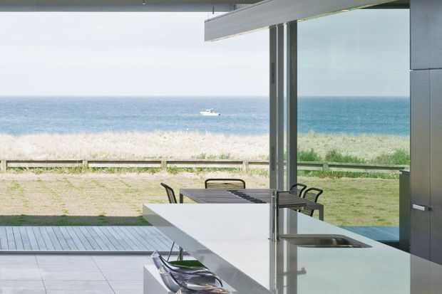 A contemporary open-plan kitchen opening directly onto a deck, with a long white island and integrated sink, dark fixed cabinetry, and an outdoor dining setting visible beyond the full-height glazing. The space overlooks a sandy beachfront and ocean, creating a bright coastal outlook.