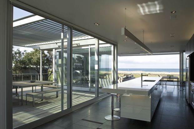 A sleek contemporary kitchen opening onto a covered outdoor dining terrace, with a large white island, dark cabinetry, grey tiled flooring, extensive sliding glass doors, and a direct view toward the beach and ocean. Linear pendant lighting and horizontal roof screening reinforce the clean modern design.