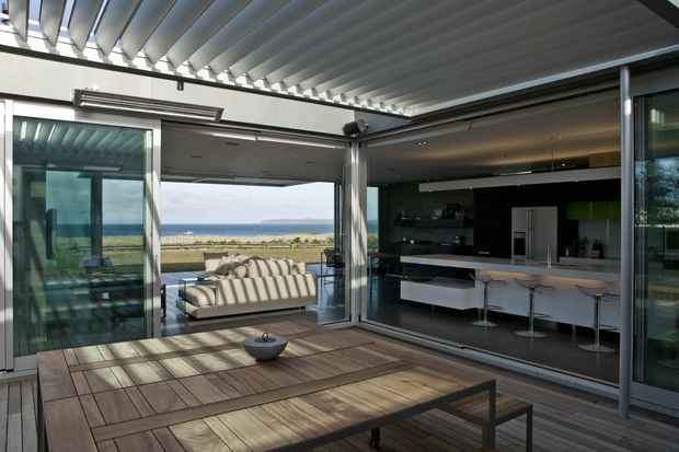 A contemporary open-plan living and outdoor entertaining space with large retractable glazed doors opening toward a sandy beachfront and ocean view. The room features a dark modern kitchen with a long island and bar stools, a light sofa, timber decking, and a covered pergola with strong linear slatted structure. Indoor and outdoor areas flow together through extensive glazing.