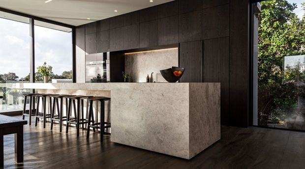 A refined contemporary kitchen featuring a large stone island with integrated seating, dark timber cabinetry, a recessed illuminated display niche, floor-to-ceiling glazing, and dark timber flooring. The open layout overlooks greenery and a residential landscape.