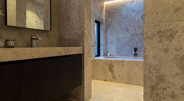 A refined contemporary bathroom clad extensively in warm beige and taupe natural stone, with a floating dark timber vanity, pale stone benchtop, wall-mounted basin tap, and large mirrored cabinet. A recessed bathing or shower area is visible beyond, framed by a narrow dark window and softly illuminated concealed strip lighting.