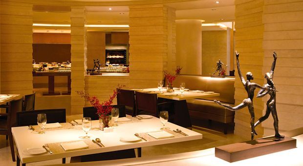 Upscale restaurant dining room with formally set tables, upholstered banquette seating, warm layered lighting, stone-clad columns and walls, and a prominent sculptural human-form artwork. An open service or kitchen area is visible in the background.