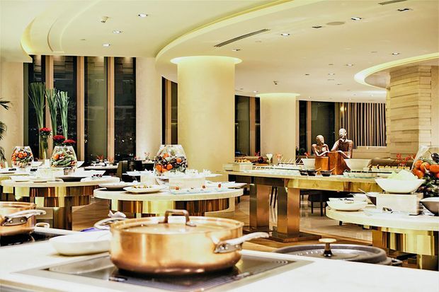 A spacious, upscale commercial buffet and dining interior with multiple circular serving islands, copper cookware, prepared food displays, and polished counters. The room features large cylindrical columns, curved cream-colored ceiling forms with recessed lighting, floor-to-ceiling dark glazing, and a sculptural curved feature wall. Staff or diners are visible in the background.