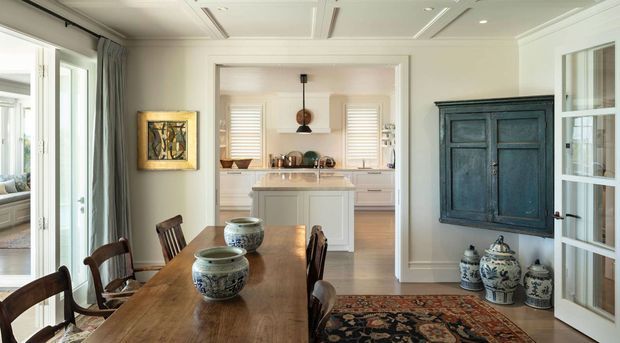 A refined dining room with a long solid-wood dining table and traditional chairs, opening directly to a bright white kitchen with a central island. The room features pale walls, detailed ceiling mouldings, tall glazed doors, grey curtains, patterned rug, decorative blue-and-white ceramics, and a dark blue built-in wall cabinet. The overall look is classic and coastal-influenced with abundant natural light.