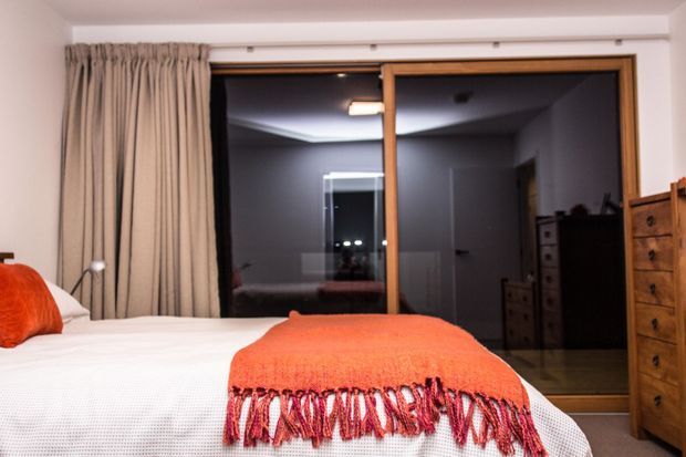 A warmly styled bedroom viewed toward a large timber-framed sliding glass door. The room contains a white bed with a vivid orange-red throw and cushion, beige full-length curtains, and a wooden chest of drawers at right. The glazing opens visually onto a dark adjoining or exterior space, with reflected room details visible in the glass. The architecture is simple and contemporary, with a neutral palette accented by orange-red textiles.