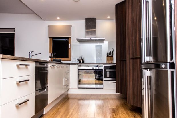 Bright contemporary U-shaped kitchen with white flat-panel cabinetry, dark timber-toned tall storage, stainless-steel appliances, a pale stone benchtop, glossy white splashback, and warm timber flooring. The compact layout includes an oven, cooktop, rangehood, dishwasher, sink, and integrated refrigeration, with recessed ceiling lights and a small window.
