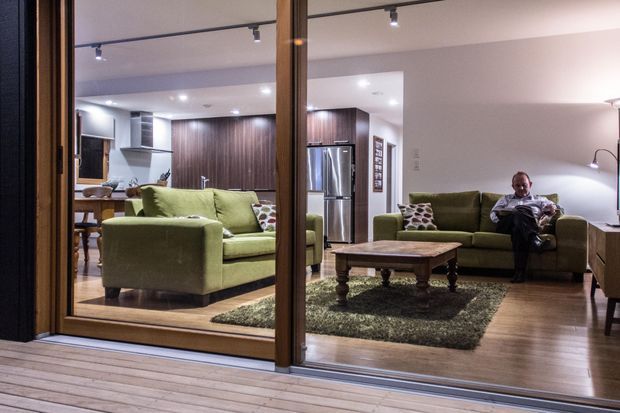Interior view through large timber-framed glazing into an open-plan living room, with two green sofas, a carved wooden coffee table, dark textured rug, timber flooring, and a compact kitchen with dark wood cabinetry and stainless-steel appliances in the background. Recessed and track lighting create a warm, contemporary atmosphere. A timber deck is visible in the foreground.