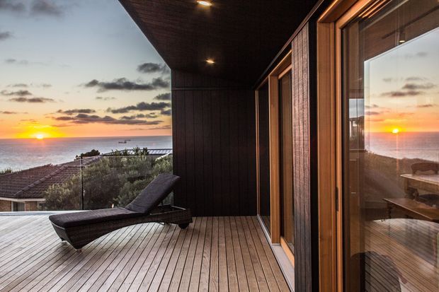 A timber-clad coastal terrace or balcony overlooking the ocean at sunset. The spacious deck has linear timber decking, dark vertical wall cladding and ceiling, recessed lighting, large timber-framed glazed doors, and a reclining outdoor lounge chair. The glazing reflects an interior dining or living area, while neighbouring rooftops and coastal vegetation are visible beyond the balustrade.