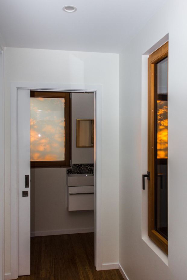 A narrow, clean-lined interior hallway or passage with white painted walls and ceiling, dark timber flooring, and warm timber-framed windows. An open doorway leads into a compact bathroom or vanity area with a wall-mounted white cabinet, dark mosaic tile splashback, and reflective window glass. Recessed ceiling lighting and simple black hardware provide restrained contemporary detailing.
