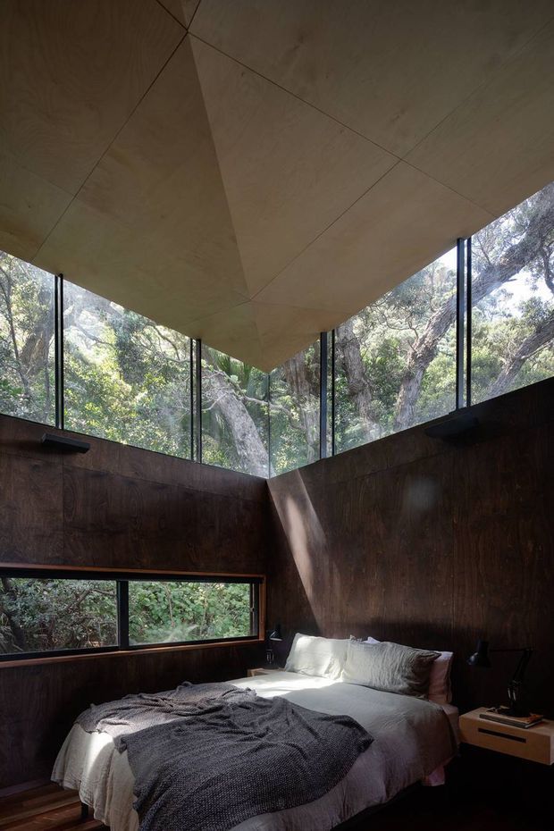 A dramatic bedroom in a forest setting, featuring dark timber wall panelling, extensive floor-to-ceiling glazing, and a sculptural plywood-lined ceiling with angular geometry. A double bed sits beneath the glazing, with simple bedside tables and dark lighting fixtures. Dense native woodland is visible outside.