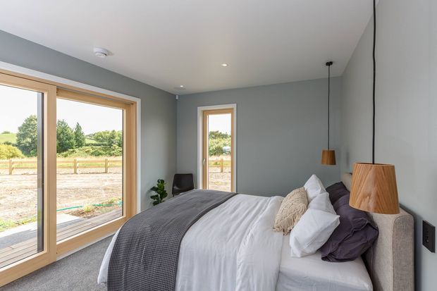 A bright contemporary bedroom with a double bed dressed in white bedding, a charcoal grey throw, and muted purple cushions. The room has soft grey walls, a white ceiling, light timber-framed floor-to-ceiling glazing and a narrow matching window, with timber pendant lights and views onto a rural landscape.