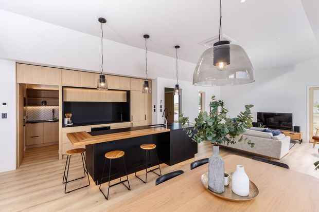 Open-plan contemporary kitchen, dining and living area with light timber cabinetry and flooring, a large black island with timber benchtop, integrated television joinery, pendant lighting, and a dining table in the foreground. The space is bright and refined, with a neutral palette and indoor plants.