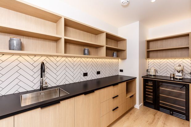 A contemporary kitchen with light timber cabinetry and open shelving, a black benchtop, stainless-steel sink and tapware, and a white herringbone tile splashback. Integrated under-shelf lighting illuminates the work area, while a separate black wine-storage unit and appliance station sit to the right. Light timber flooring and a neutral palette create a warm, refined look.