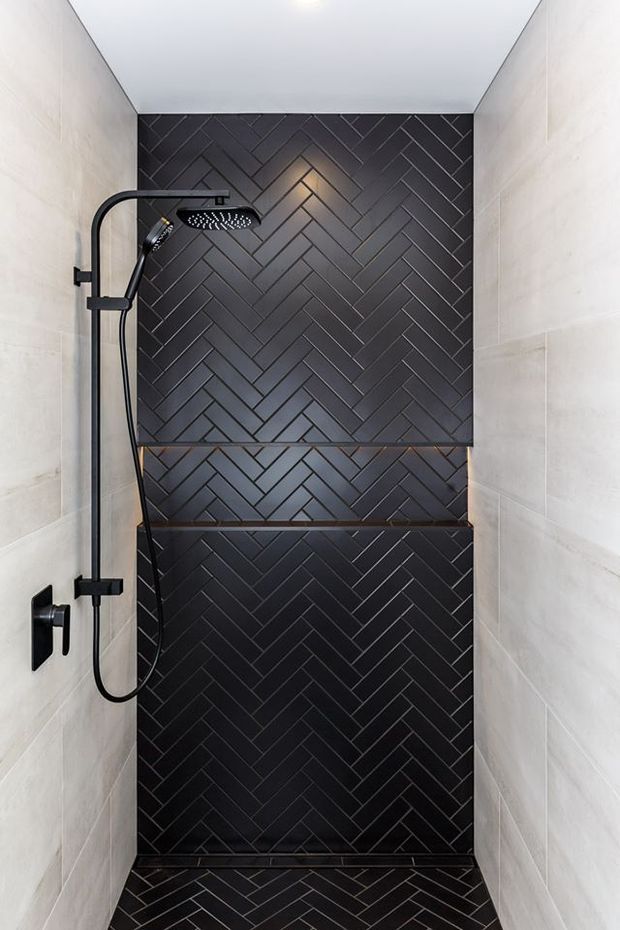 A compact modern walk-in shower with a matte black rain shower and handheld fixture. The rear wall and built-in ledges are clad in dark charcoal herringbone tile, contrasted with light stone-look wall tiles on both sides. Black fixtures and minimal detailing create a refined, high-contrast bathroom interior.