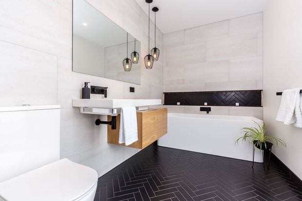 Contemporary bathroom featuring a freestanding white bathtub, wall-mounted timber vanity with white basin, large frameless mirror, white tiled walls, and dark herringbone floor and backsplash tiles. Pendant lights hang above the vanity, with minimal black fixtures and a small indoor plant.