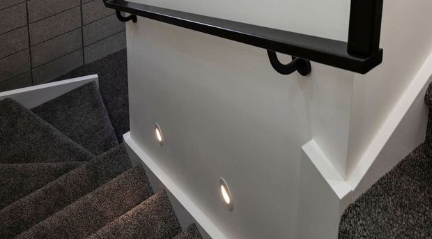 A modern carpeted stairway viewed from above, featuring dark grey textured carpet, white plasterboard walls, recessed wall lights, and a black metal handrail with brackets.