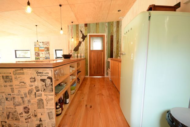 Warm, compact retro-inspired kitchen with extensive timber finishes, open shelving, a large pale mint-green freestanding refrigerator, and a timber breakfast bar or island faced with vintage newspaper-print panels. The room includes a patterned feature wall, timber cabinetry, pendant lighting, and a timber door, creating a distinctive eclectic vintage character.