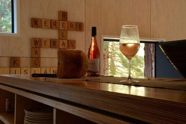 Warm, intimate interior detail showing a timber-clad wall with a wooden letter-tile display, a layered timber kitchen or bar counter, open shelving, a bottle, wine glass, and a bread loaf. A window and interior opening bring in views of surrounding trees, while strong sunlight creates a cozy, rustic atmosphere.