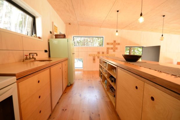 Warm, compact timber-lined kitchen with extensive plywood cabinetry, a central island, integrated open shelving, a retro pale-green refrigerator, exposed plumbing at the sink, and pendant lights. Large windows provide views to surrounding trees, while the timber floor, walls, and ceiling create a cohesive natural interior.