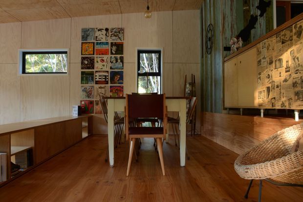 A compact, warm timber-lined interior combining a dining area and living space. The room features plywood wall and ceiling panels, timber flooring, a simple rectangular dining table with mid-century wooden chairs, low built-in timber cabinetry, a woven lounge chair, narrow windows, and an eclectic wall display of artwork and printed material. A patterned tiled or papered feature wall adds visual interest behind the built-in joinery.