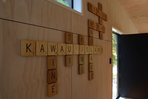 Interior wall clad in warm timber plywood, featuring a decorative arrangement of wooden Scrabble-style letter tiles spelling “Kawa Island” and other words. A narrow high window and open dark-framed doorway bring in natural light, with greenery visible outside.