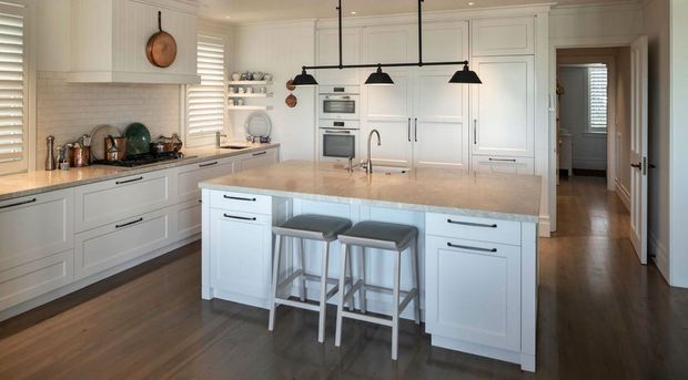 Bright, spacious white kitchen with extensive shaker-style cabinetry, a large central island with a pale stone benchtop and two stools, integrated appliances, dark timber flooring, horizontal window shutters, and black pendant lighting. The kitchen opens to a hallway and adjoining spaces.