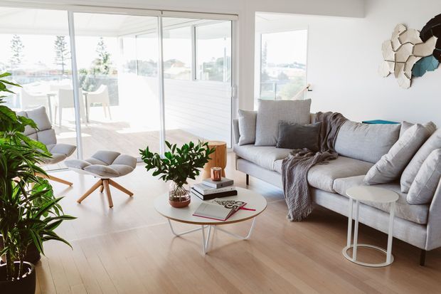 Bright, airy contemporary living room with pale timber flooring, a large light-grey sectional sofa, white walls, and extensive sliding glass doors opening to a glazed outdoor space. The room includes a white-and-wood coffee table, nesting side tables, a lounge chair with ottoman, indoor plants, and muted neutral furnishings. Coastal views and a dining area are visible beyond the glazing.