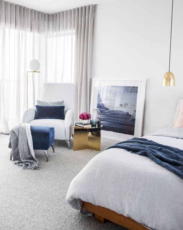 Bright, refined bedroom with a soft neutral palette and navy accents. The room features a large upholstered bed, layered white bedding, navy throw and cushions, a pale armchair with matching ottoman, full-height sheer and grey drapery, light carpet, a slim gold side table, pendant light, and large framed artwork leaning against the wall.