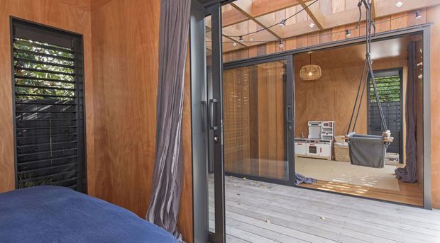 View from a bedroom through dark-framed glazed doors into a warm timber-lined indoor-outdoor living or relaxation space. The room features extensive timber wall and ceiling finishes, louvred windows, grey curtains, pale timber decking or flooring, a hanging chair, and a small utility or kitchen appliance area visible beyond.