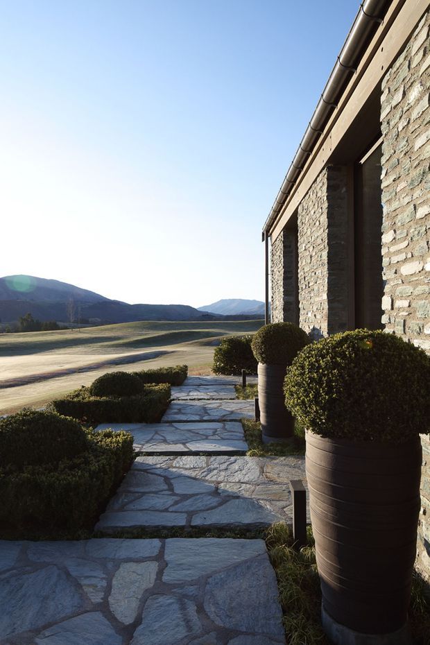 Exterior architectural view of a stone-clad building beside a landscaped stone pathway. The facade features rough natural stone walls, dark recessed openings, and a pitched roof with metal gutters. Large rounded topiary shrubs in dark planters line the irregular stone paving, with an expansive rural mountain landscape visible beyond.