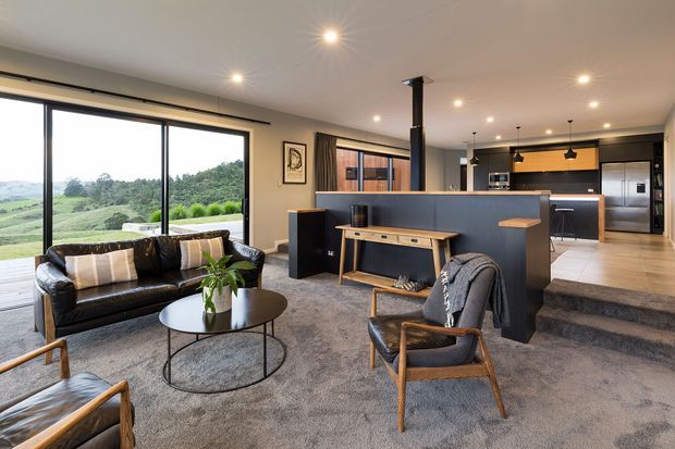 Open-plan contemporary living and kitchen area with expansive floor-to-ceiling glazing overlooking a green rural landscape. The room features a dark leather sofa and armchairs, round coffee table, textured carpet, timber accents, a dark kitchen island with timber breakfast ledge, pendant lighting, and warm recessed ceiling lights. A short set of steps connects the living zone to the kitchen.