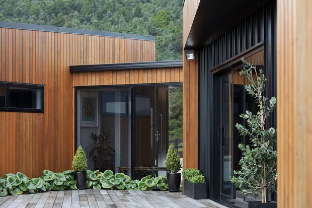 Contemporary timber-clad building arranged around a landscaped courtyard, with vertical warm wood siding, dark metal-framed glazing, black roof edges, and a timber-paved terrace. Dense greenery and a forested hillside form the background.