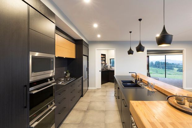 Contemporary kitchen with dark full-height cabinetry, integrated stainless-steel appliances, black tiled splashback, timber overhead cabinetry, and a large central island with a warm timber benchtop. Three black pendant lights hang above the island, while large glazing provides views to a green rural landscape.