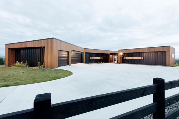 A wide exterior view of a low, angular contemporary residence with a long horizontal composition. The facade combines warm vertical timber cladding with dark ribbed wall panels and recessed garage or entry openings. A broad pale concrete driveway curves across the foreground, bordered by a black timber fence and sparse landscaping. The overcast sky and simple geometric forms create a restrained, modern architectural presentation.