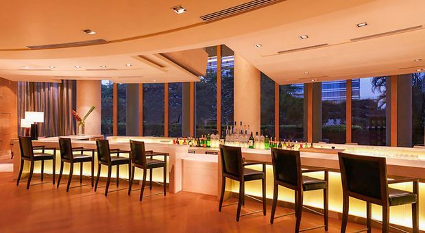 A warmly lit, contemporary bar and dining area with a long illuminated stone or light-coloured counter, numerous bar stools, dining tables and chairs, recessed ceiling lighting, timber flooring, and floor-to-ceiling windows overlooking greenery. Bottles and glassware are arranged along the bar, creating an upscale hospitality-style interior.