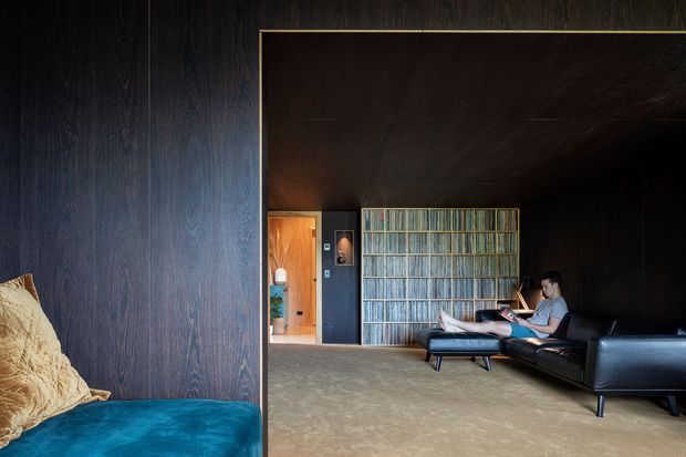 Dark, sophisticated interior lounge viewed from an adjacent seating area. The room features dark timber wall and ceiling lining, a broad warm-grey carpeted floor, a black leather sectional sofa and chaise, and a blue upholstered foreground seat. A full-height textured translucent or ribbed glass shelving partition divides the lounge from a timber-lined entry or adjoining space. The atmosphere is moody and refined, with strong clean-lined contemporary detailing.