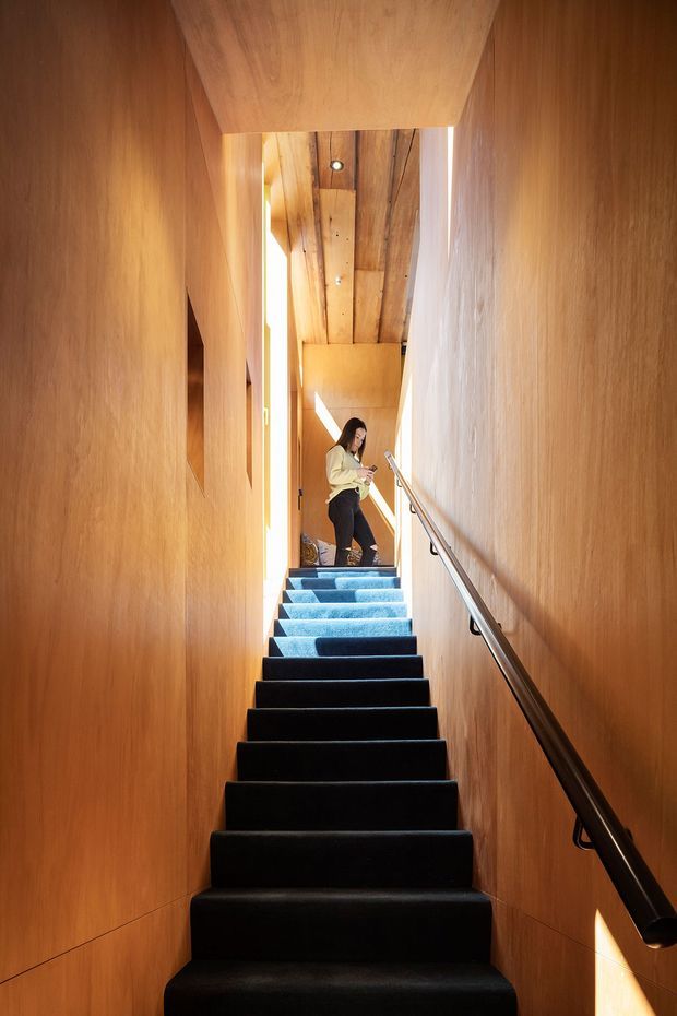 A narrow, vertically framed interior stairway with dark carpeted steps ascending toward a bright landing. The walls and ceiling are clad in warm timber, with a simple metal handrail along the right side. A person stands at the upper landing, and daylight enters from the side, creating strong highlights and shadows.