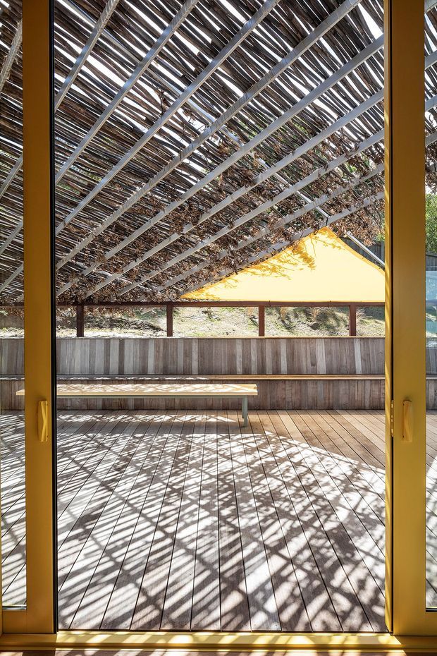 View from an interior through large yellow-framed glazed doors onto a timber deck and built-in bench seating. A substantial timber pergola with closely spaced slatted screening creates strong patterned shadows, while a bright yellow triangular shade or roof form is visible beyond. The scene is sunlit and naturally integrated with surrounding vegetation.