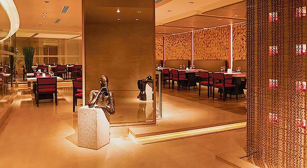 A warmly lit, upscale restaurant interior with multiple dining areas, red upholstered dining chairs, dark tables, patterned feature walls, polished light flooring, large mirrors, recessed ceiling lighting, and a decorative hanging chain partition. Sculptural artwork is displayed near the mirrored divider.