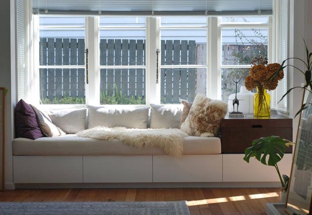Bright, cozy sitting area with a built-in white window-seat sofa beneath a bank of tall multi-pane windows. Neutral cushions, layered cream throws, a dark wood sideboard, indoor plants, and warm timber flooring create a calm, softly styled interior.