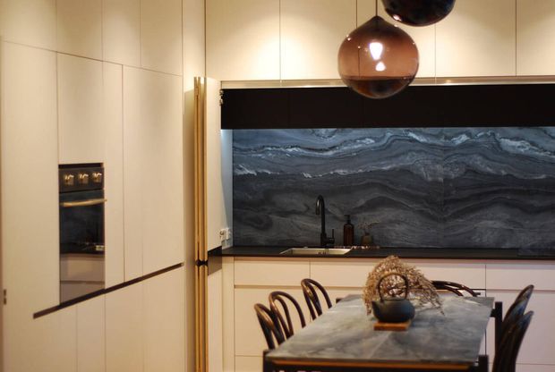 A refined contemporary kitchen and dining area with full-height cream cabinetry, integrated appliances, a dramatic dark veined stone splashback, brass-toned vertical detailing, and a matching stone dining table. Sculptural amber pendant lights and curved dark dining chairs add warmth and visual contrast.