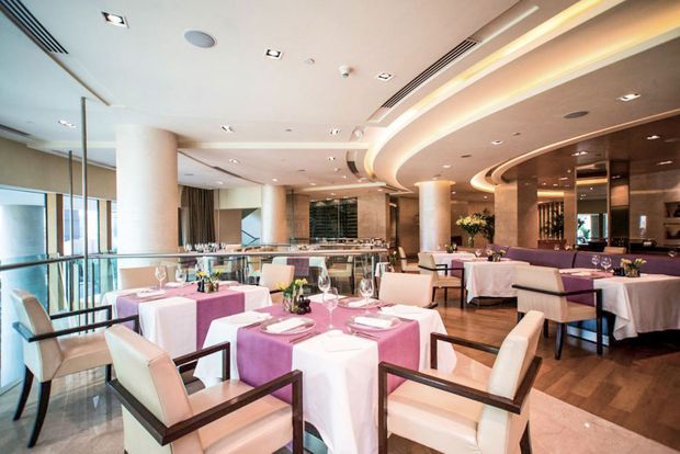 Upscale contemporary restaurant dining room with neatly dressed tables, upholstered armchairs, a bar or service counter at the rear, extensive glazing, and curved recessed ceiling details with integrated lighting. The interior uses warm wood flooring, pale upholstery, dark timber accents, and pink-purple table linens.