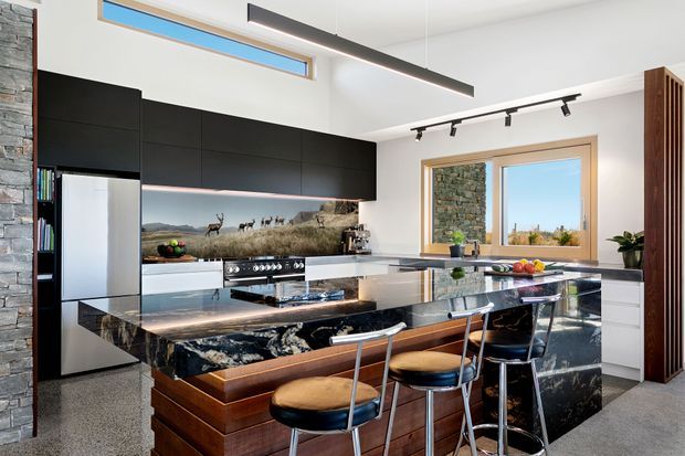 Contemporary kitchen with a large veined stone island and waterfall ends, dark flat-panel cabinetry, integrated stainless-steel refrigerator, and timber drawer fronts. Three bar stools line the island. A stone-clad wall, timber-framed windows, high clerestory glazing, and linear track and pendant lighting contribute to the bright, refined interior. A panoramic landscape backsplash is visible behind the cooktop.