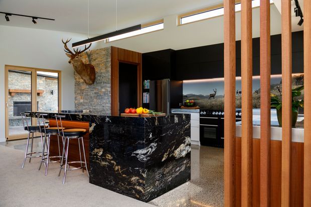 A dramatic contemporary kitchen and dining area with a large black marble waterfall island, dark cabinetry, integrated stainless-steel appliances, and a timber slat divider. A stone feature wall, clerestory windows, track lighting, and a compact dining table complete the space. The kitchen opens toward a view of mountainous landscape through the glazing.